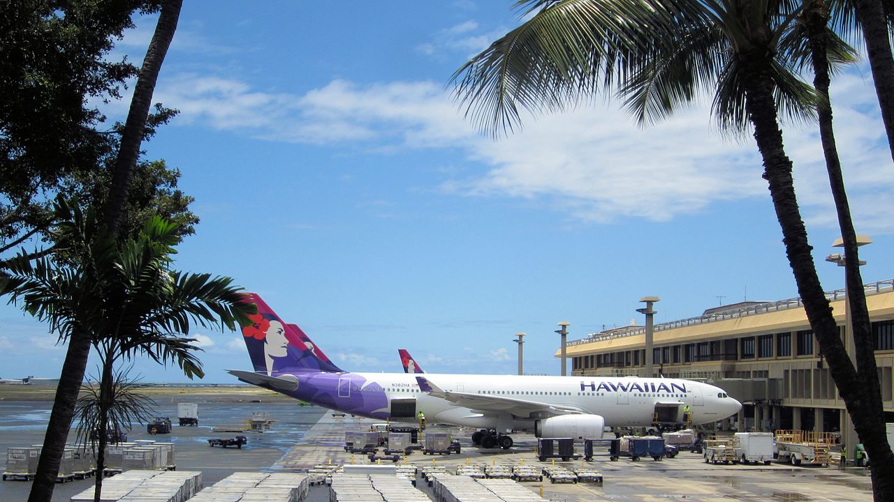 A Hawaiian Airlines Airbus parked at a tree lined terminal.