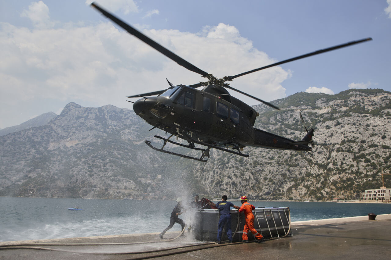 A Montenegro Air Force Bell 412 EPi helicopter hovers over a Bambi Bucket for firefighting.