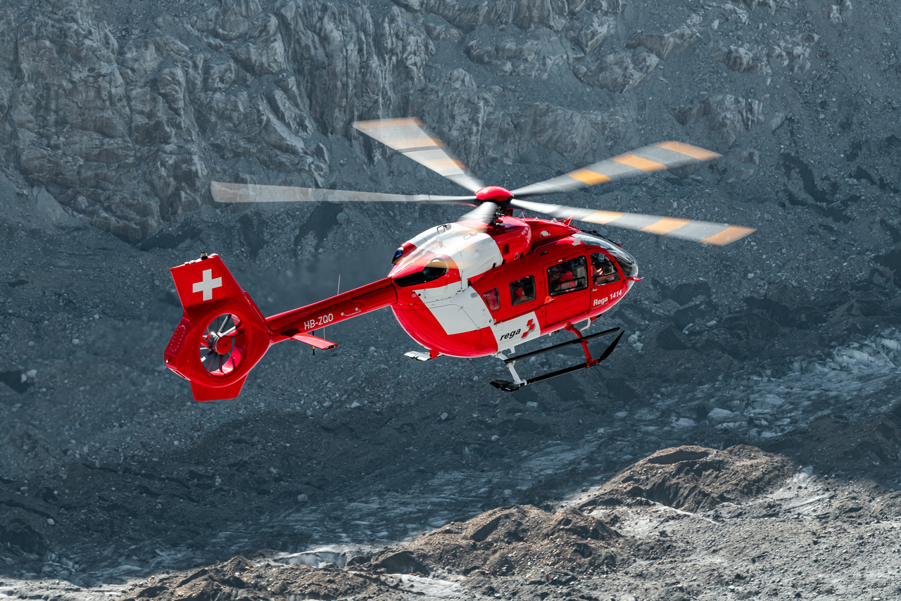 A Rega Swiss Air Rescue Airbus H145 helicopter over mountainous terrain.