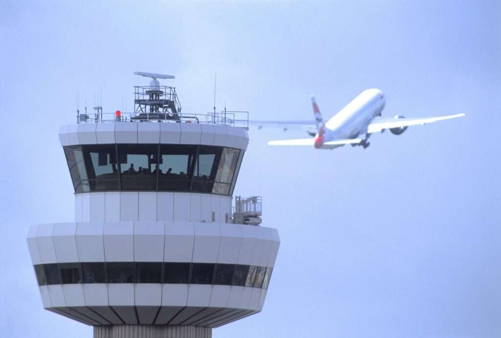 Gatwick Airport control tower with aircraft in flight in background.